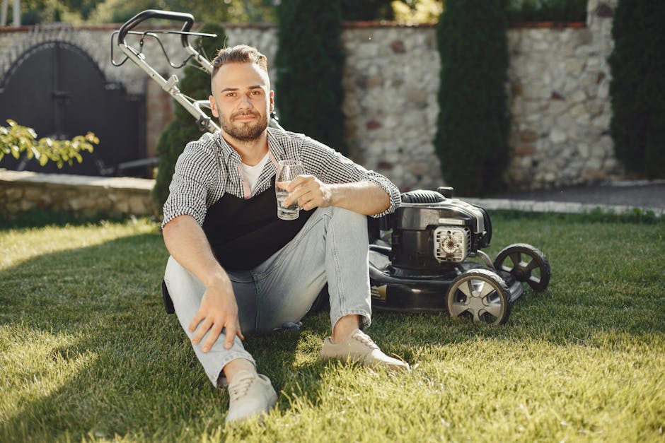 A man with a beard and short hair, dressed in a checkered shirt, black apron, and light-colored jeans, is sitting on a well-maintained grassy lawn in a garden. He is holding a glass of water in his ri