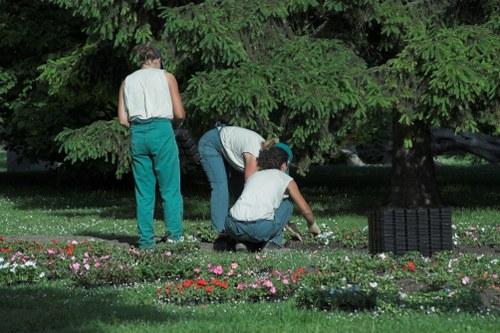 A landscaped garden area featuring a well-maintained grassy lawn bordered by flower beds with pink, red, and white blooms. In the background, large mature trees with lush green foliage provide shade and structure to the outdoor space. Three individuals are engaged in gardening activities: one standing while observing, and two others crouched down, actively tending to the flower beds, possibly planting or weeding. The garden is adjacent to a paved or stone pathway, and a black garden waste bin is visible on the right side of the image. Natural daylight illuminates the scene, suggesting a clear weather day, with the vibrant colors of the flowers and greenery contrasting with the neutral tones of the clothing and garden features, illustrating a peaceful outdoor environment suitable for gardening services in Staines.
